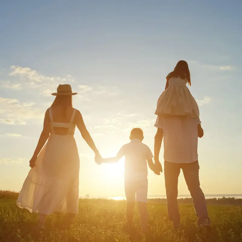 Happy family enjoys walking together on green meadow. Father carries daughter on shoulders holding boy hand walking on lawn against blue sky at sunset